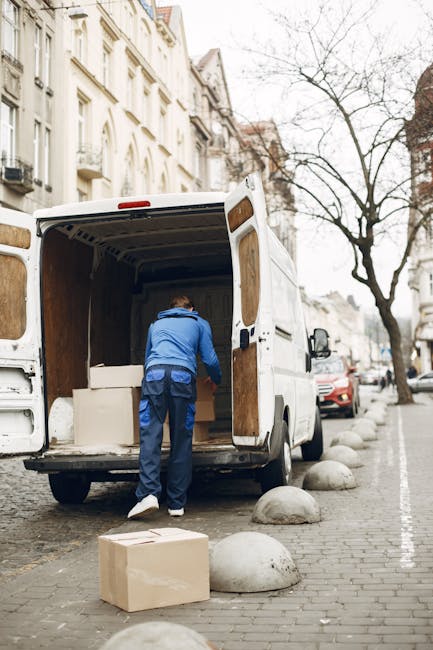 Two men wearing dark clothing and baseball caps are engaged in home relocation tasks outside a residential property. One man is pushing a hand truck loaded with large cardboard boxes toward a white va