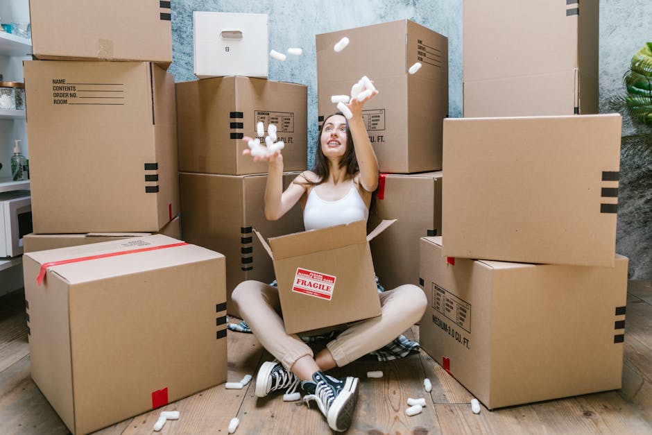 A person lying on their back behind a pile of brown cardboard moving boxes and packing supplies inside a room with light wood flooring and a white paneled door in the background. The boxes are various