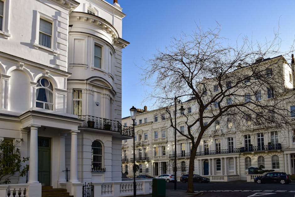 A clear view of a street in a residential area during daytime, featuring a row of white Victorian-style terraced houses with ornate architectural details such as bay windows, decorative molding, and iron balconies. In the foreground, a leafless tree with extensive branches extends towards a bright blue sky. On the pavement, a black lamppost is positioned near the curb, while parked cars are visible along the street. In the background, a van from Man and Van Maida Hill is parked close to one of the properties, with a furniture trolley and packing materials, including cardboard boxes, plastic wrapping, and moving blankets, placed near the entrance. The scene captures the process of home relocation, with furniture and packing materials prepared for transportation, illustrating a typical urban house move in Maida Hill.