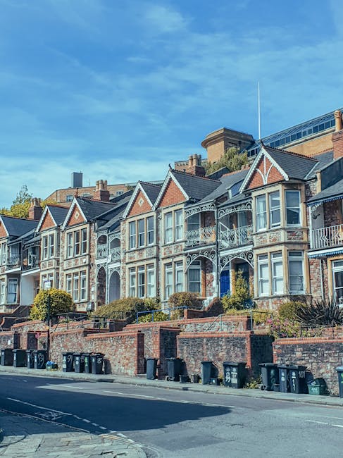 Photograph of a row of elegant white Victorian-style terraced houses with ornate architectural details, including decorative window surrounds, balconies with wrought iron railings, and classical columns at ground level, located along Maida Hill in Westbourne Park. The buildings are illuminated by natural daylight, casting soft shadows on the facade, and the clear blue sky is visible above. A black street lamp and some leafless trees are also seen in the foreground. This setting depicts a residential street prepared for a home relocation or furniture transport, with the pavement in front of the homes ready for loading and unloading activities, possibly by Man and Van Maida Hill during their house removals and packing process.