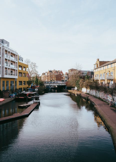 A view of a narrow urban canal lined with green trees and low-rise brick and stone buildings on the right side, with a paved walkway alongside the water. Several boats are moored along the canal's edg